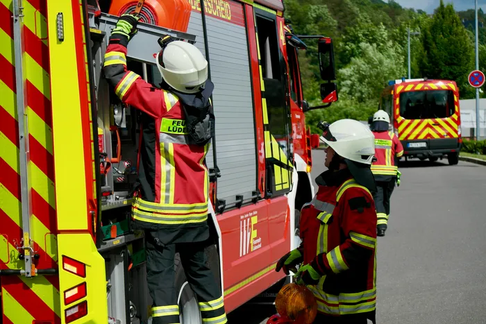 Feuerwehrleute in roter und schwarzer Einsatzkleidung mit Helmen arbeiten an der Seite eines roten Feuerwehrautos. Im Hintergrund steht ein weiterer Feuerwehrauto und Bäume.