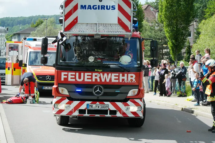 Roter Feuerwehrauto der Marke Magirus mit der Aufschrift „FEUERWEHR Brögge“ fährt auf eine Gruppe von Zuschauern zu. Im Hintergrund sind weitere Einsatzfahrzeuge und Menschen zu sehen.