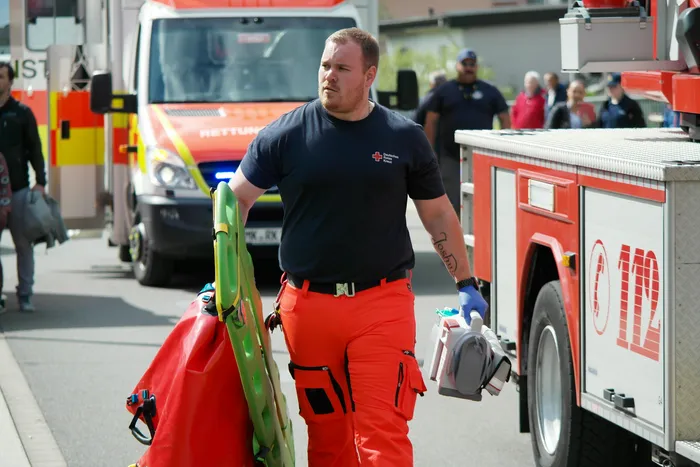 Ein Rettungssanitäter in schwarzem Polohemd und orangefarbener Hose trägt eine Trage und einen medizinischen Koffer vor einem Rettungswagen. Im Hintergrund sind weitere Einsatzkräfte und Fahrzeuge zu sehen.