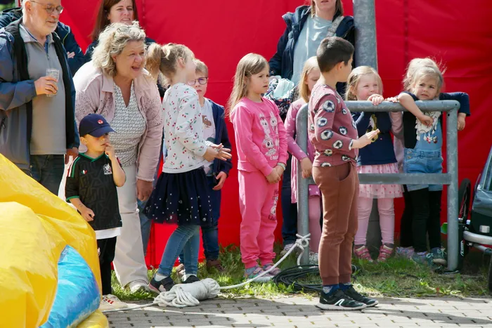 Eine Gruppe von Kindern und Erwachsenen steht an einem Zaun, einige schauen in Richtung einer gelben Hüpfburg. Im Hintergrund ist ein roter Stoffbanner zu sehen.