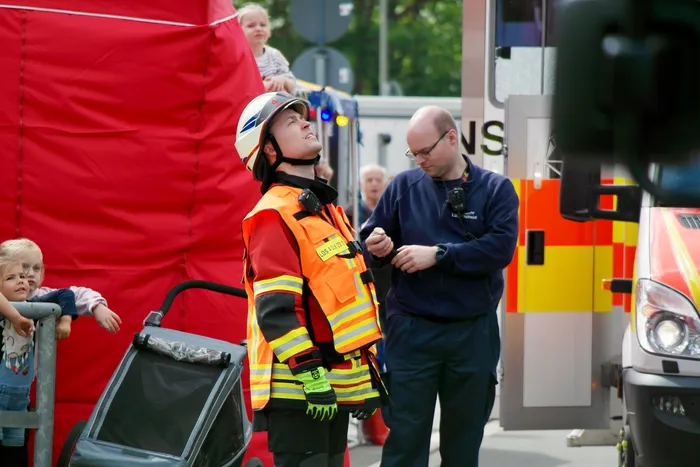 Ein Feuerwehrmann in voller Montur steht neben einem Polizisten, der in sein Funkgerät spricht. Im Hintergrund ist ein roter Lastwagen und ein kleines Kind zu sehen, das aus einem Fenster schaut.