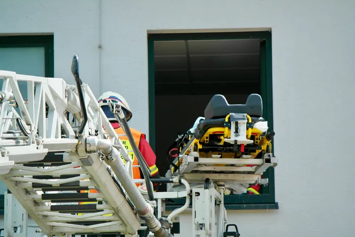 Rettungskräfte positionieren eine Trage aus einem Rettungswagen an einem Fenster eines Gebäudes, während ein Feuerwehrmann im Hintergrund steht. Die Aufnahme zeigt einen Teil einer Drehleiter.