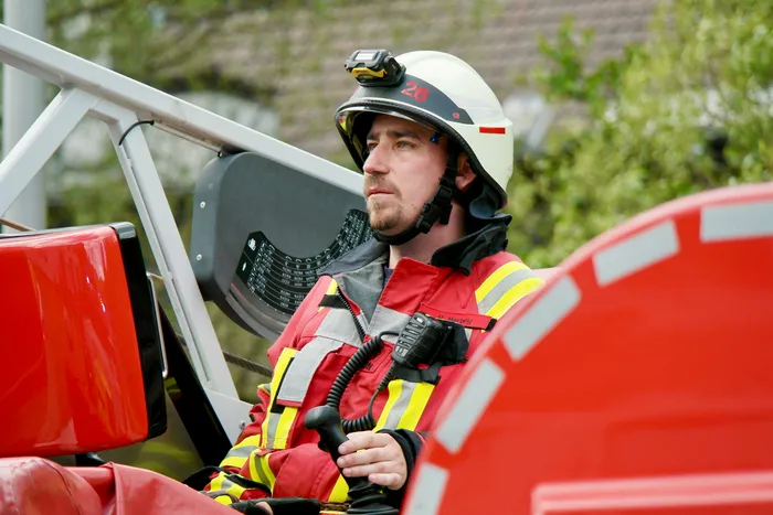 Feuerwehrmann mit Helm und Funkgerät sitzt konzentriert am Steuer eines roten Einsatzfahrzeugs. Im Hintergrund ist ein verschwommener Busch zu sehen.