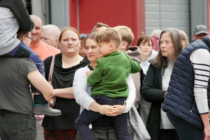 Eine Frau trägt ein Kind auf ihren Schultern, umgeben von weiteren Menschen im Freien. Im Hintergrund sind Gebäude und weitere Besucher zu sehen.