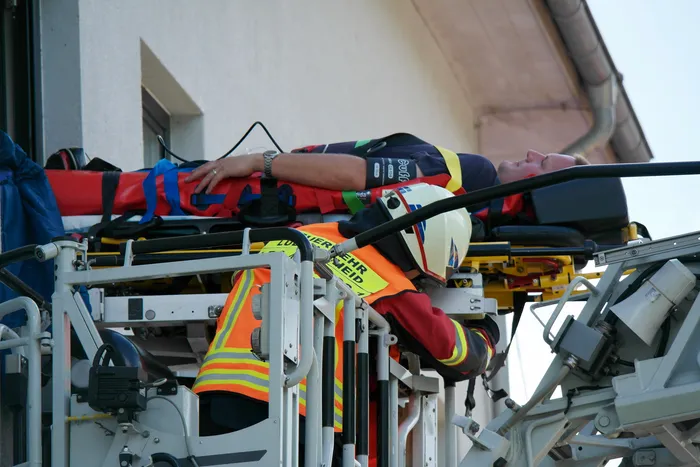 Ein Rettungssanitäter behandelt eine Person auf einer Trage, die von Feuerwehrleuten aus einem Gebäude gerettet wurde. Im Hintergrund ist ein rotes Gebäude und Ausrüstung der Feuerwehr zu sehen.