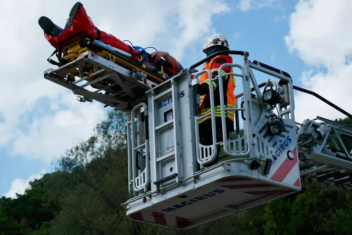 Ein Feuerwehrmann in voller Montur sichert eine Trage auf einer Hebebühne. Im Hintergrund sind Bäume und ein blauer Himmel mit Wolken zu sehen.