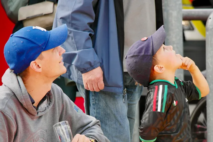 Ein Mann und ein Kind schauen aufmerksam nach oben, beide tragen Baseballkappen. Im Hintergrund sind weitere Personen und ein Geländer zu sehen.