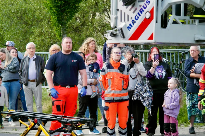Menschen beobachten eine simulierte Rettungsaktion mit einem Rettungswagen und Einsatzkräften in orangefarbenen Westen. Eine Frau fotografiert mit einer Kamera, während andere Zuschauer mit ihren Handys filmen.