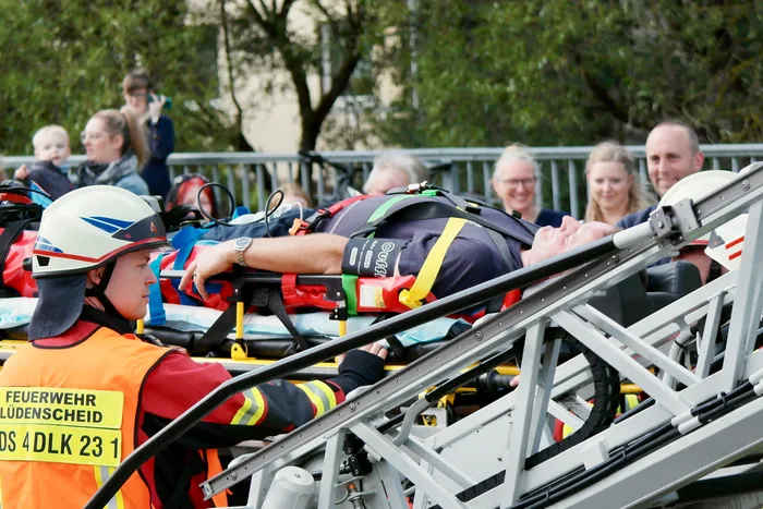 Feuerwehrleute sichern eine Person auf einer Trage, die mit einer Hebebühne abgeseilt wird. Im Hintergrund stehen Zuschauer und beobachten die Übung.