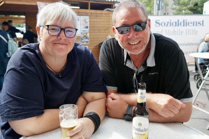 Zwei lächelnde Menschen sitzen an einem Tisch mit Biergläsern und Flaschen vor einem Stand mit dem Aufdruck “Sozialdienst und Mobile Hilfe”. Im Hintergrund sind weitere Menschen und ein Rollstuhl zu sehen.