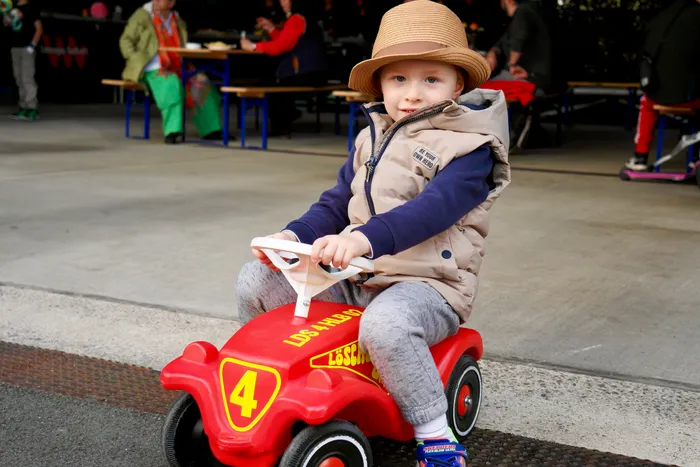 Ein kleiner Junge mit Strohhut sitzt auf einem roten Spielzeugauto und hält das Lenkrad fest. Im Hintergrund sitzen und stehen Menschen an Tischen in einem Freiluftbereich.