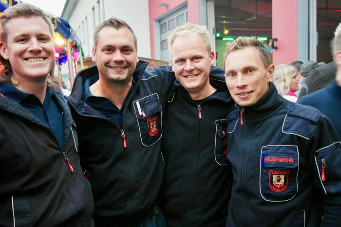 Vier Feuerwehrleute in dunkelblauen Jacken mit Aufnähern lächeln in die Kamera. Im Hintergrund sind Menschen und ein Festzelt zu sehen.