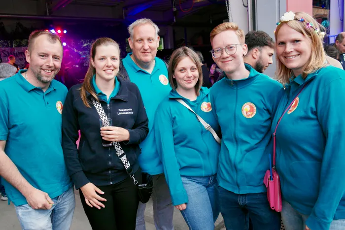 Eine Gruppe von Feuerwehrleuten in türkisfarbenen Poloshirts und Jacken steht lächelnd zusammen. Im Hintergrund sind weitere Menschen und ein festlich geschmückter Bereich zu sehen.
