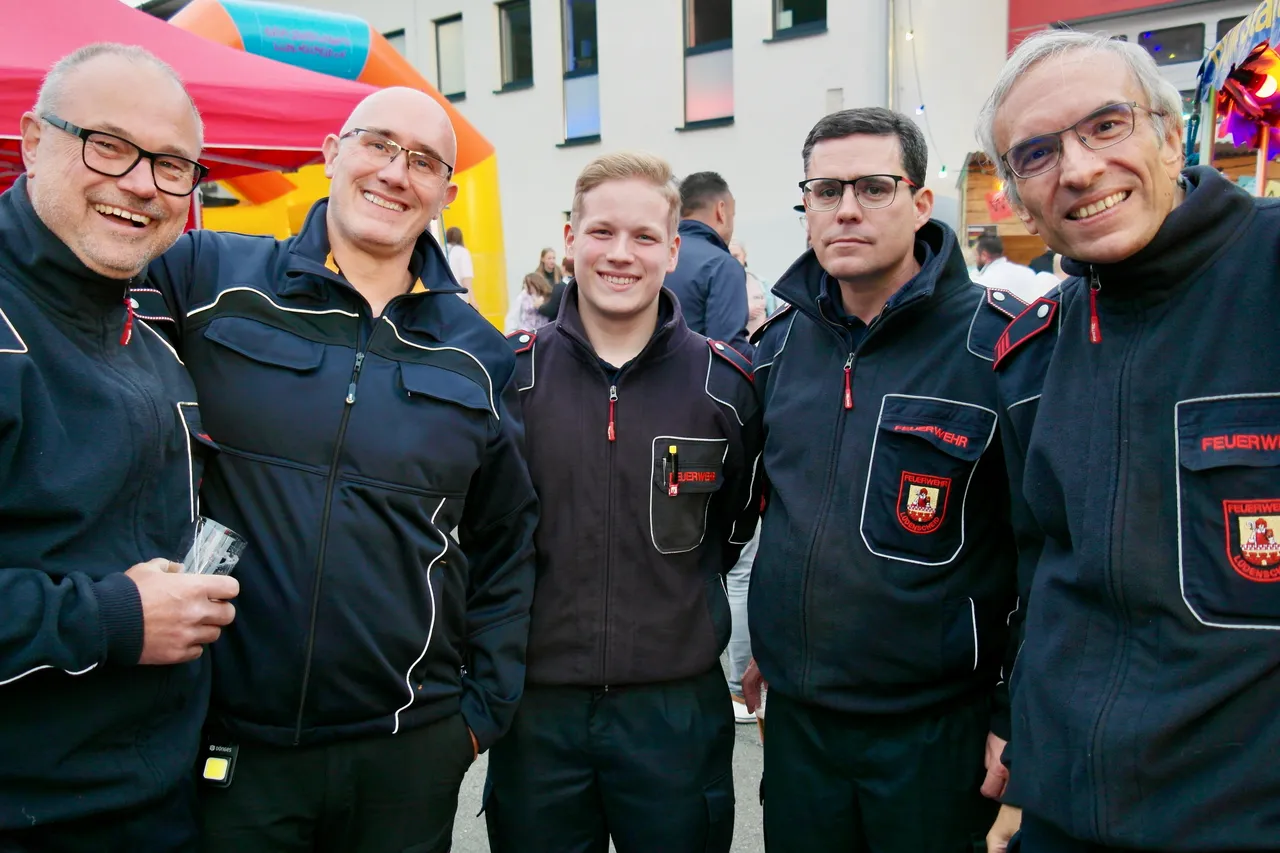 Fünf Feuerwehrleute in schwarzer Uniform mit roten Akzenten stehen lächelnd vor einer bunten Kulisse aus Festzelten und Attraktionen. Sie tragen Abzeichen und wirken stolz auf das Fest des Löschzugs Brügge.