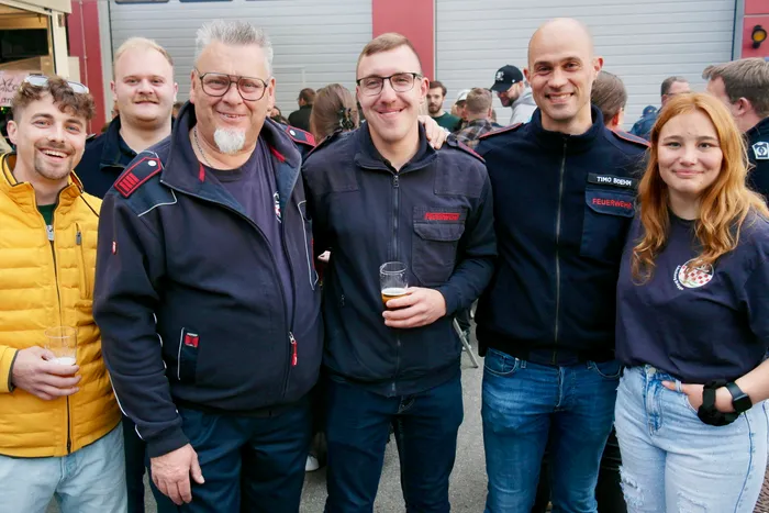 Fünf lächelnde Menschen stehen eng zusammen, einige halten Getränke in der Hand. Zwei Männer tragen Uniformen der Freiwilligen Feuerwehr, während die anderen zivil gekleidet sind. Im Hintergrund ist ein rotes Feuerwehrauto zu sehen.