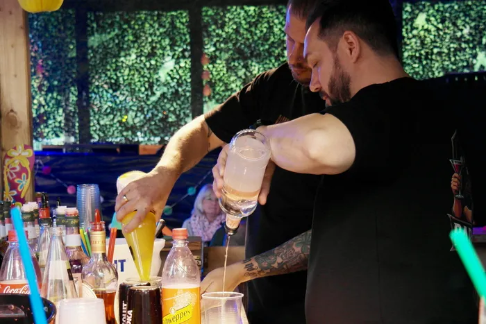 Ein Mann mit Tattoos schenkt ein Getränk in ein Glas. Im Hintergrund sind weitere Getränkeflaschen und Personen zu sehen.