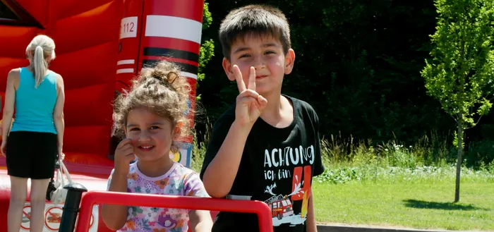Zwei Kinder posieren vor einer roten Hüpfburg und einem roten Feuerwehrauto. Der Junge macht ein Friedenszeichen, das Mädchen sitzt im Feuerwehrauto und lächelt.