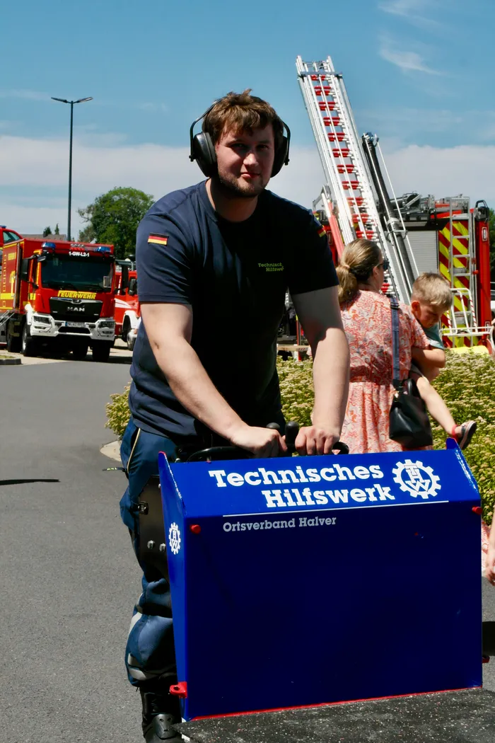 Ein junger Mann trägt eine große, blaue Tafel mit dem Aufdruck „Technisches Hilfswerk Ortsverband Halver“ und fährt auf einem Roller. Im Hintergrund ist ein rotes Feuerwehrauto und eine weitere Person zu sehen.