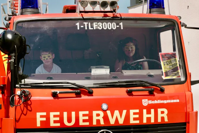 Zwei Kinder sitzen im Führerhaus eines roten Feuerwehrautos, beide mit bemalten Gesichtern. Das Fahrzeug trägt den Schriftzug 