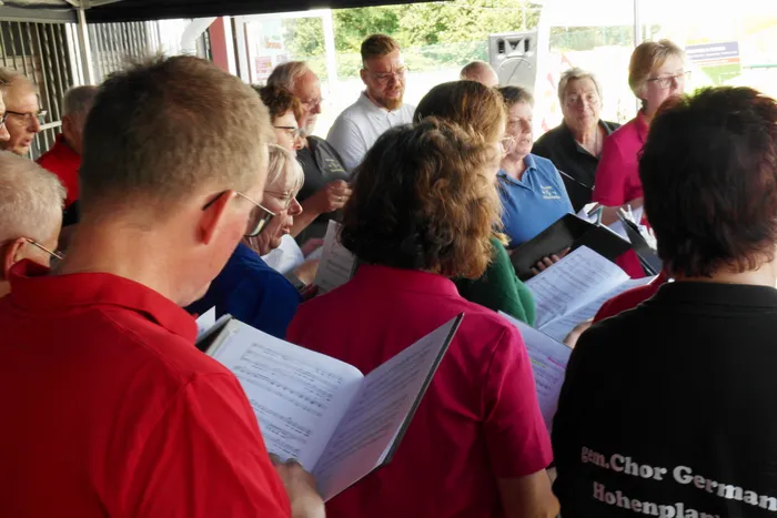 Chor-Mitglieder stehen im Freien dicht gedrängt zusammen und halten Notenblätter in den Händen. Einige singen oder lesen die Blätter, im Hintergrund sind weitere Personen und Bäume zu sehen.