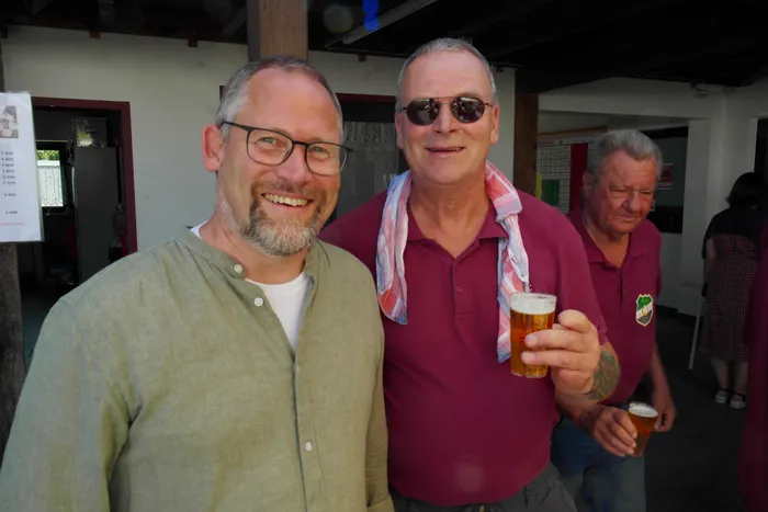 Zwei lächelnde Männer im Vordergrund, einer mit Bierglas. Im Hintergrund ein weiterer Mann und Personen an einem Sommerfest.
