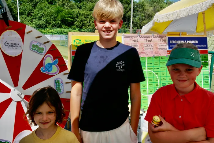 Drei Kinder stehen vor bunten Festständen und Werbeplakaten auf einem Sommerfest. Ein Junge mit blauem T-Shirt lächelt in die Kamera, während ein anderes Kind mit rotem Polohemd und ein Mädchen mit blonden Haaren daneben stehen.