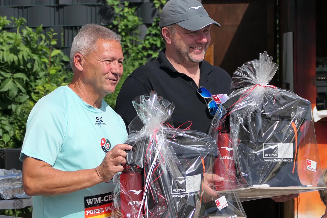 Zwei Männer halten jeweils mehrere mit roten Bändern verzierte Geschenkpakete, im Hintergrund ein Gebäude und Grünpflanzen.