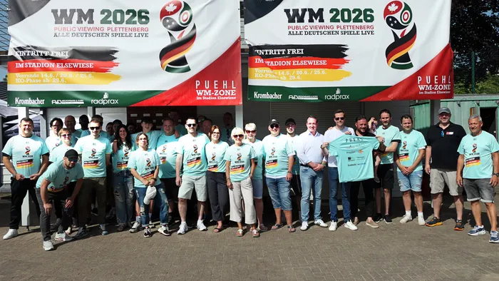 Eine Gruppe von etwa 30 Personen in hellblauen T-Shirts mit dem WM-Logo posiert vor einem Werbebanner für die Public Viewing-Veranstaltung zur Fußball-Weltmeisterschaft 2026 in Plettenberg.