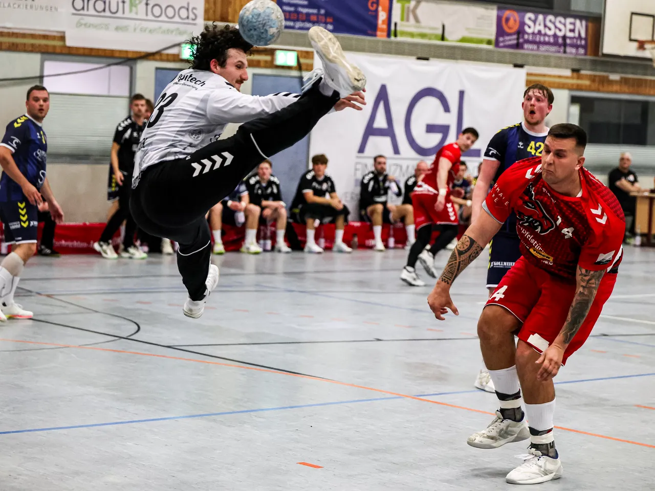 Ein Handballspieler in weiß-schwarzer Kleidung springt hoch in der Luft, um einen Ball zu erreichen, während andere Spieler auf dem Spielfeld stehen.