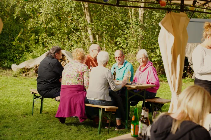 Eine Gruppe von Menschen sitzt an einem Holztisch unter einer Überdachung im Freien und unterhält sich.