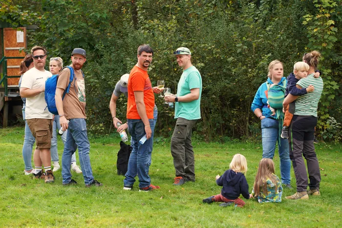 Menschen stehen bei einem Kartoffelfest auf einer Wiese vor Bäumen, einige halten Getränke, während Kinder im Vordergrund auf dem Gras spielen.