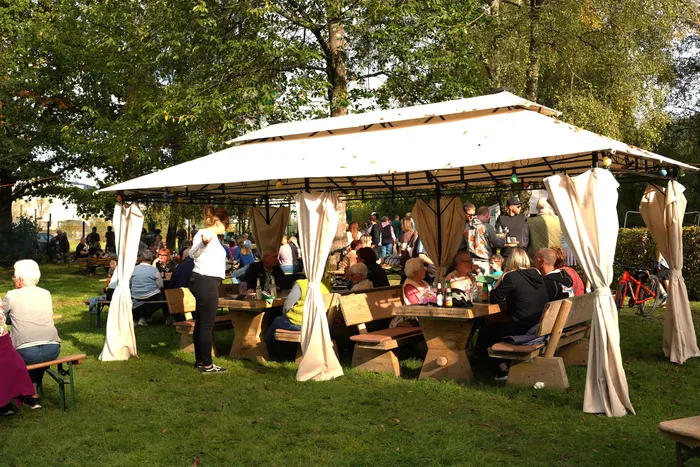 Menschen sitzen an Holztischen unter einem großen weißen Pavillon bei einem Kartoffelfest im Freien.