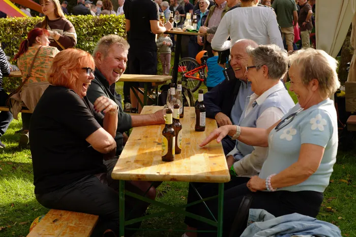 Eine Gruppe von Menschen sitzt lachend an einem Holztisch im Freien und trinkt Bier.