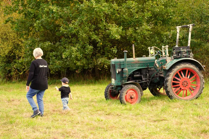 Eine Frau und ein Kind gehen auf einer Wiese zu einem alten grünen Traktor mit roten Rädern.
