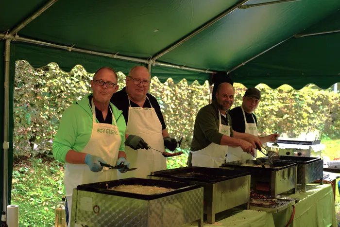 Vier Männer in Schürzen bereiten unter einem grünen Zelt Speisen auf großen Grillplatten zu.