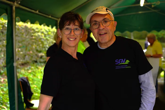 Ein lächelnder Mann mit weißer Kappe und eine Frau stehen unter einem grünen Zelt; der Mann trägt ein schwarzes T-Shirt mit dem Logo des SGV Elsetal.