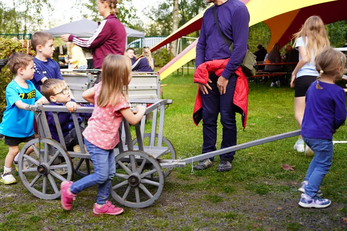 Kinder spielen mit einem hölzernen Karren auf einer Wiese vor einem gelben Festzelt beim Kartoffelfest.