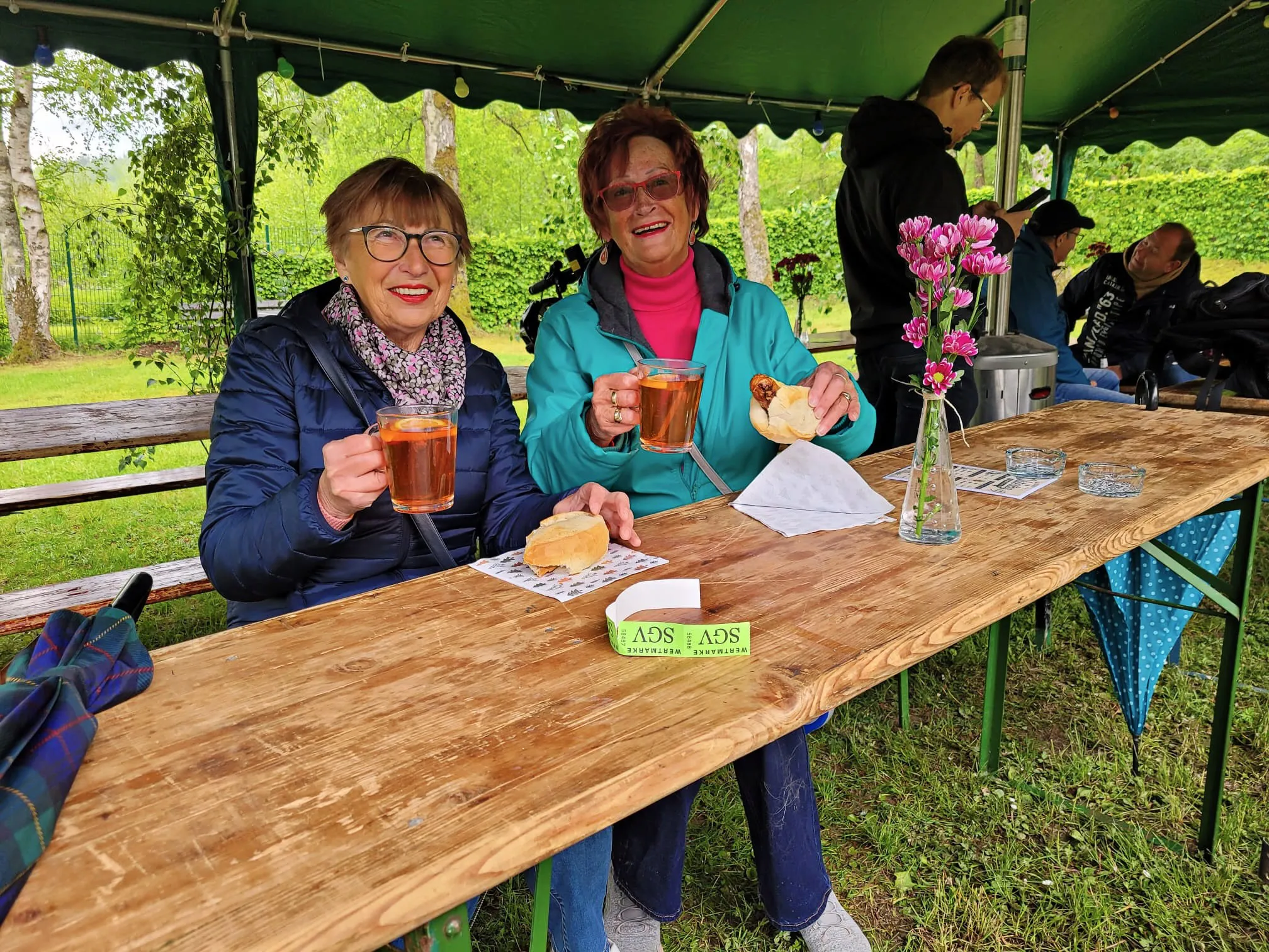 Zwei Frauen sitzen an einem gedeckten Tisch im Freien und halten jeweils ein Glas mit dunkler Flüssigkeit in der Hand. Im Hintergrund sind weitere Personen und ein Zelt zu sehen.