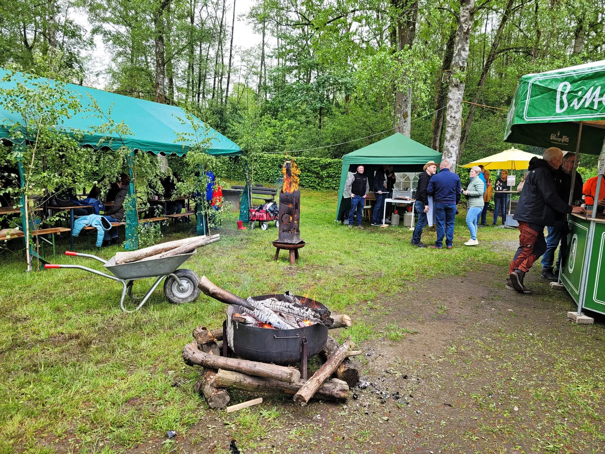Menschen stehen und sitzen im Freien vor Wanderheim und Zelten, während im Vordergrund ein Grill mit Holzkohle brennt.