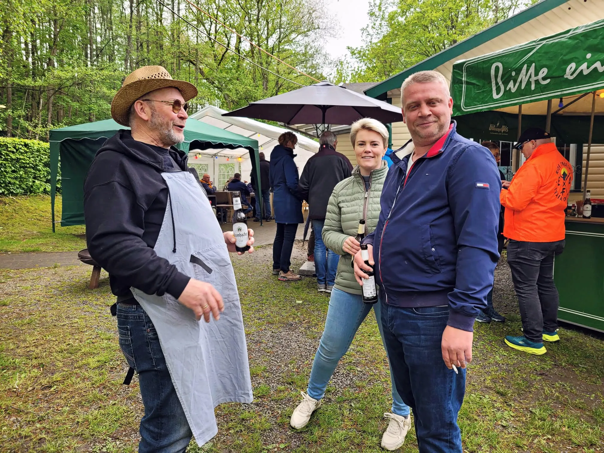 Drei Personen stehen im Freien vor einem Getränkestand, im Hintergrund weitere Besucher und Zelte eines Wandervereins.