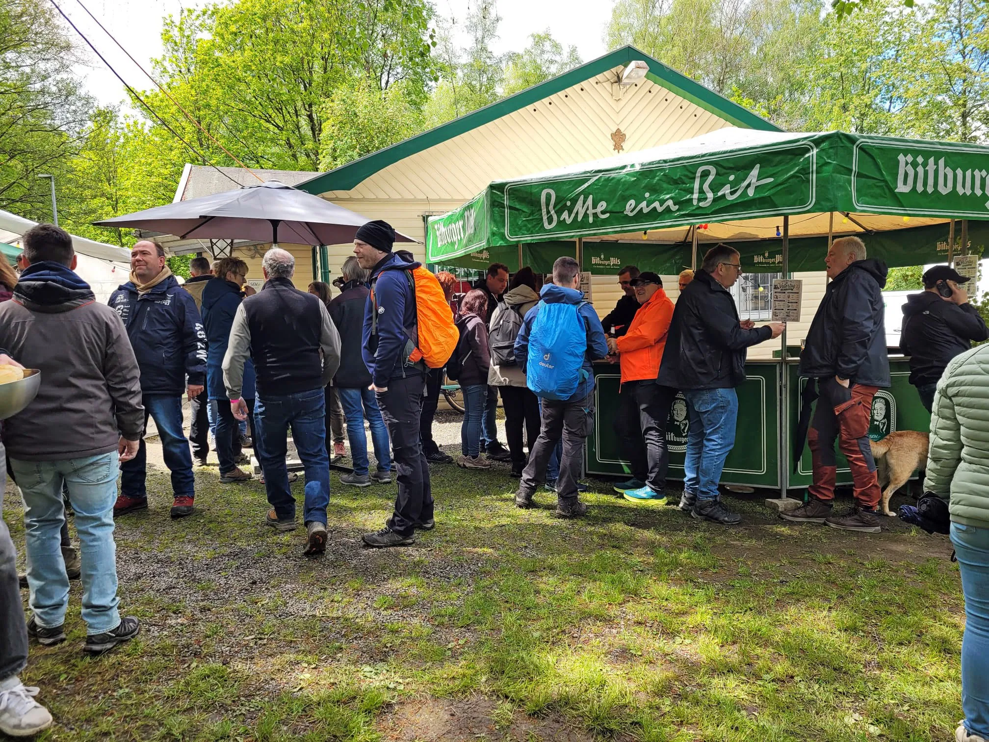 Menschen stehen an einem Imbissstand mit der Aufschrift „Bitte ein Bit“ und bestellen Getränke und Speisen. Im Hintergrund sind Bäume und weitere Besucher zu sehen.