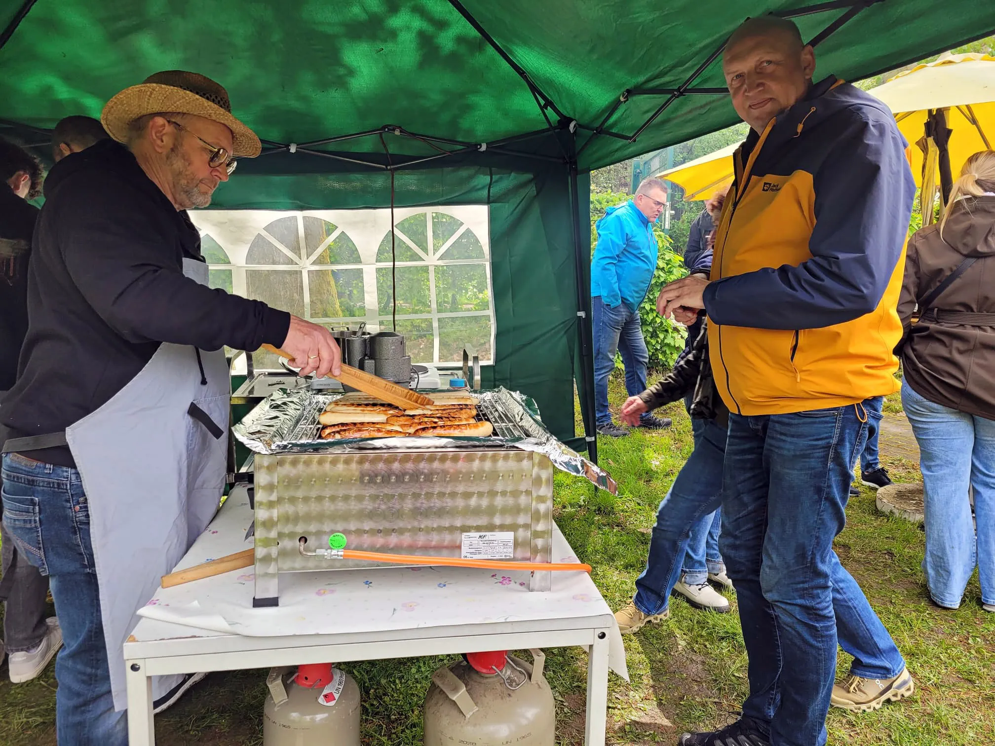 Zwei Männer stehen unter einem Pavillon an einem Grill und bereiten Essen zu, während im Hintergrund weitere Personen stehen und sich unterhalten.