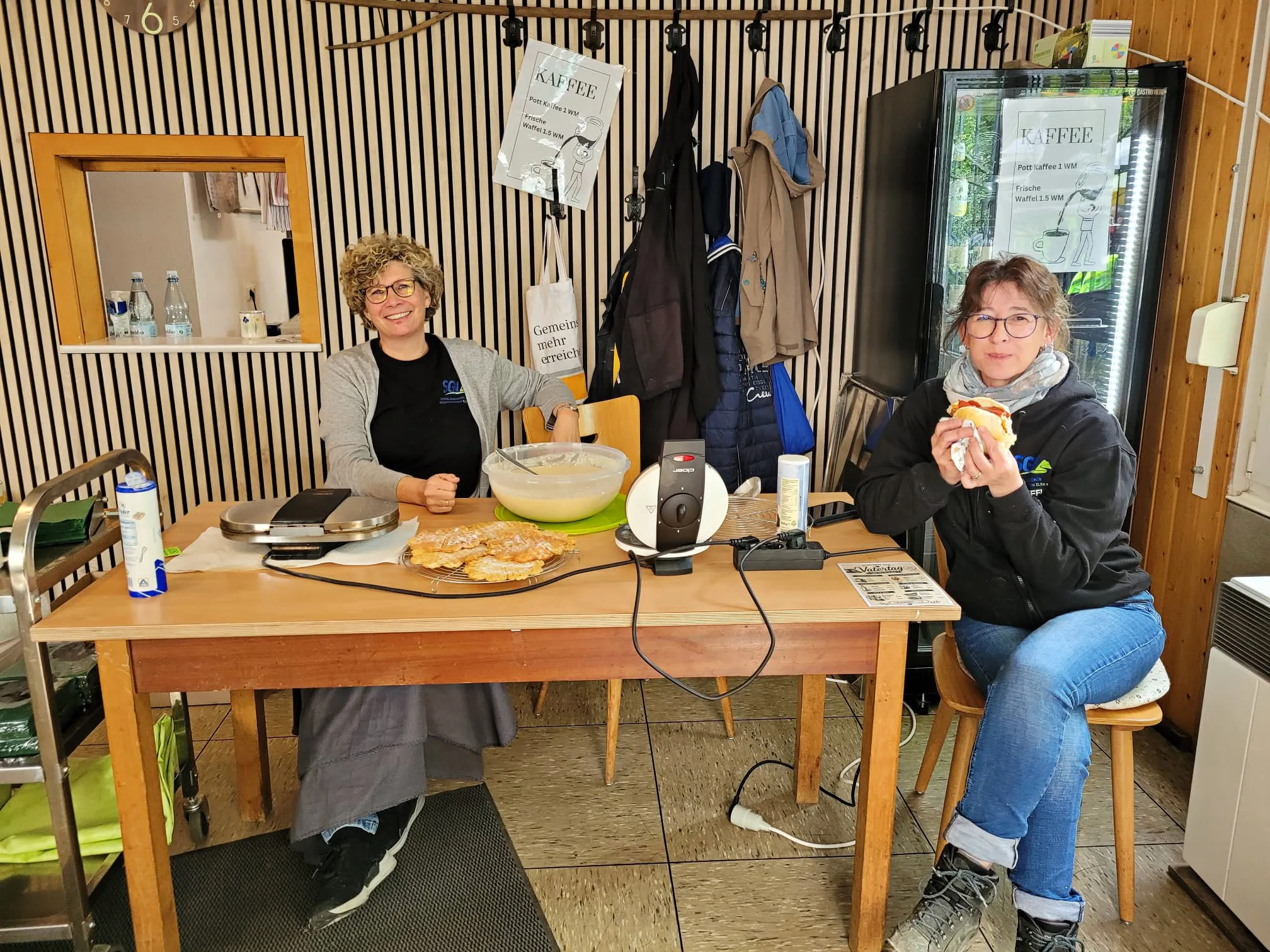 Zwei Frauen stehen hinter einem Tisch mit Gebäck und Getränken in einem Wanderheim. Im Hintergrund sind ein Kühlschrank und eine Kasse zu sehen.