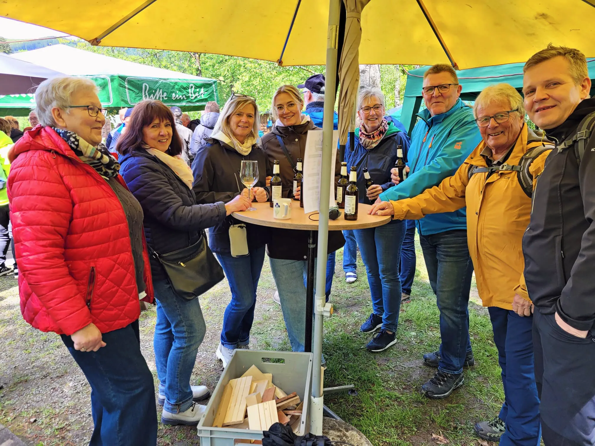 Eine Gruppe von Menschen steht um einen Tisch mit Weinflaschen unter einem gelben Zelt. Sie probieren Wein und unterhalten sich bei einer Veranstaltung des SGV Elsetal.