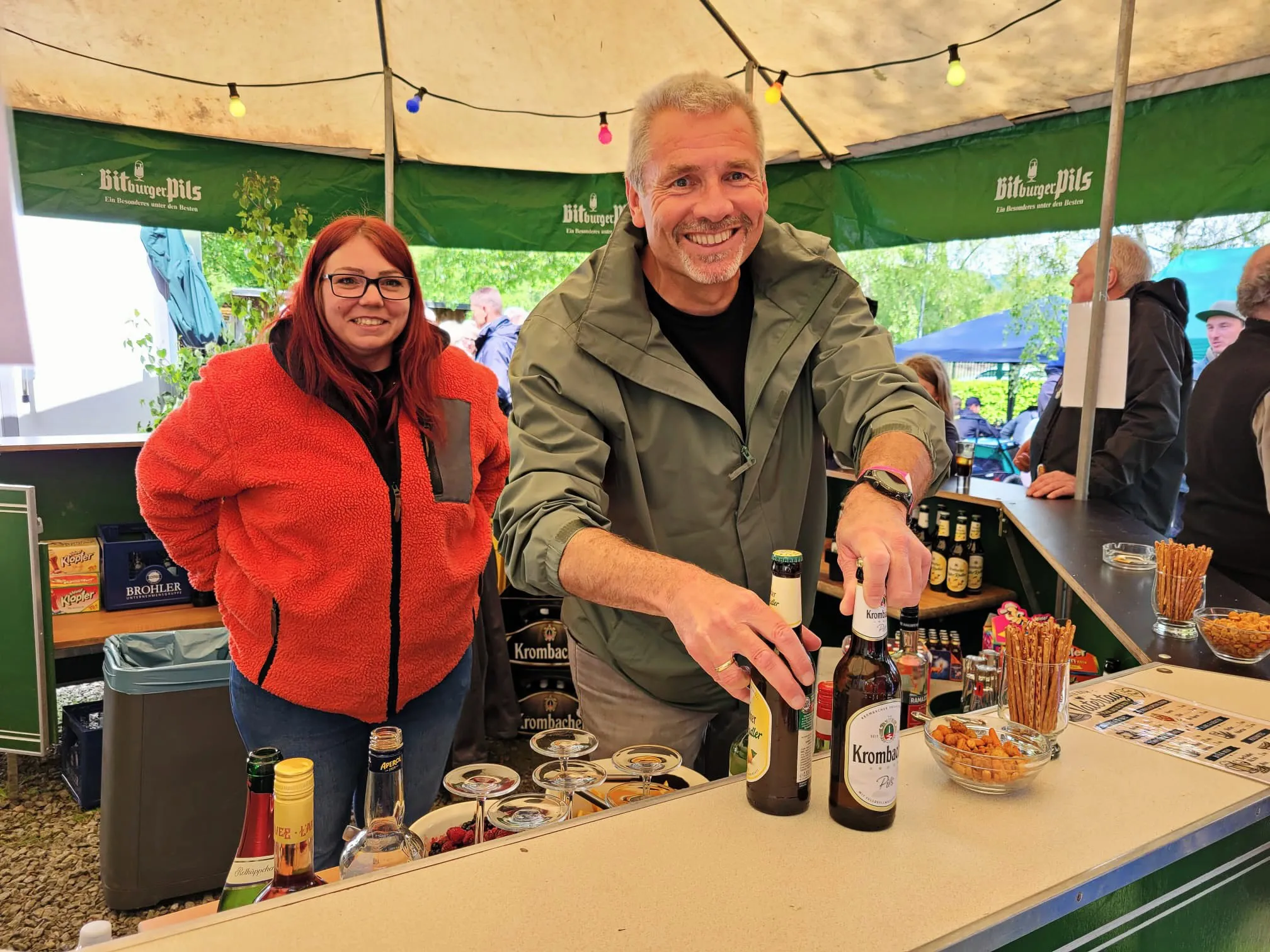 Zwei Personen stehen hinter einem Ausschankwagen mit Getränken und Snacks auf einem Volksfest. Im Hintergrund sind weitere Besucher und ein Zelt mit der Aufschrift „Bitburger Pils“ zu sehen.