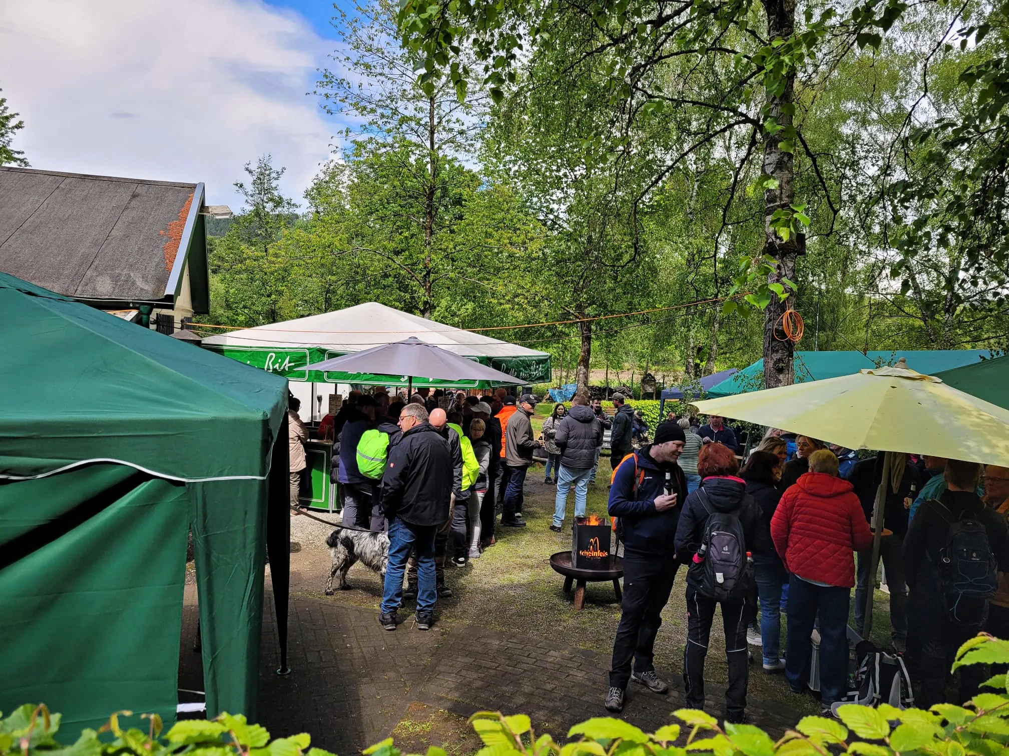 Menschengruppen stehen und unterhalten sich im Freien vor mehreren Pavillons und einem Gebäude mit rotem Dach. Im Hintergrund sind Bäume und ein blauer Himmel zu sehen.