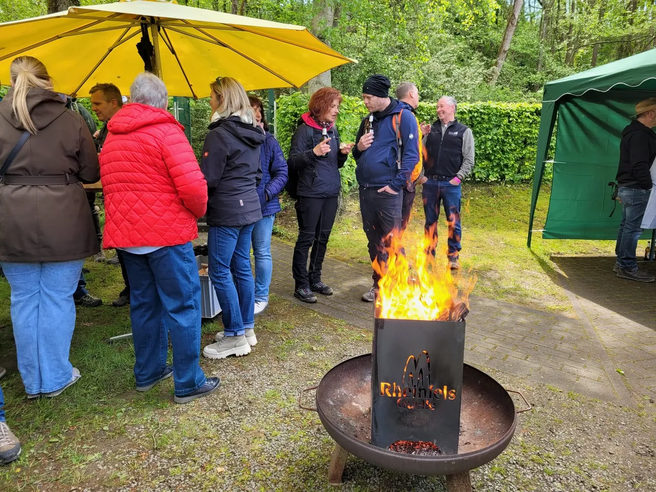 Eine Gruppe von Menschen steht um eine Feuerstelle im Freien, während ein Mann mit einem Mikrofon spricht. Im Hintergrund sind ein gelber Sonnenschirm und ein grünes Zelt zu sehen.
