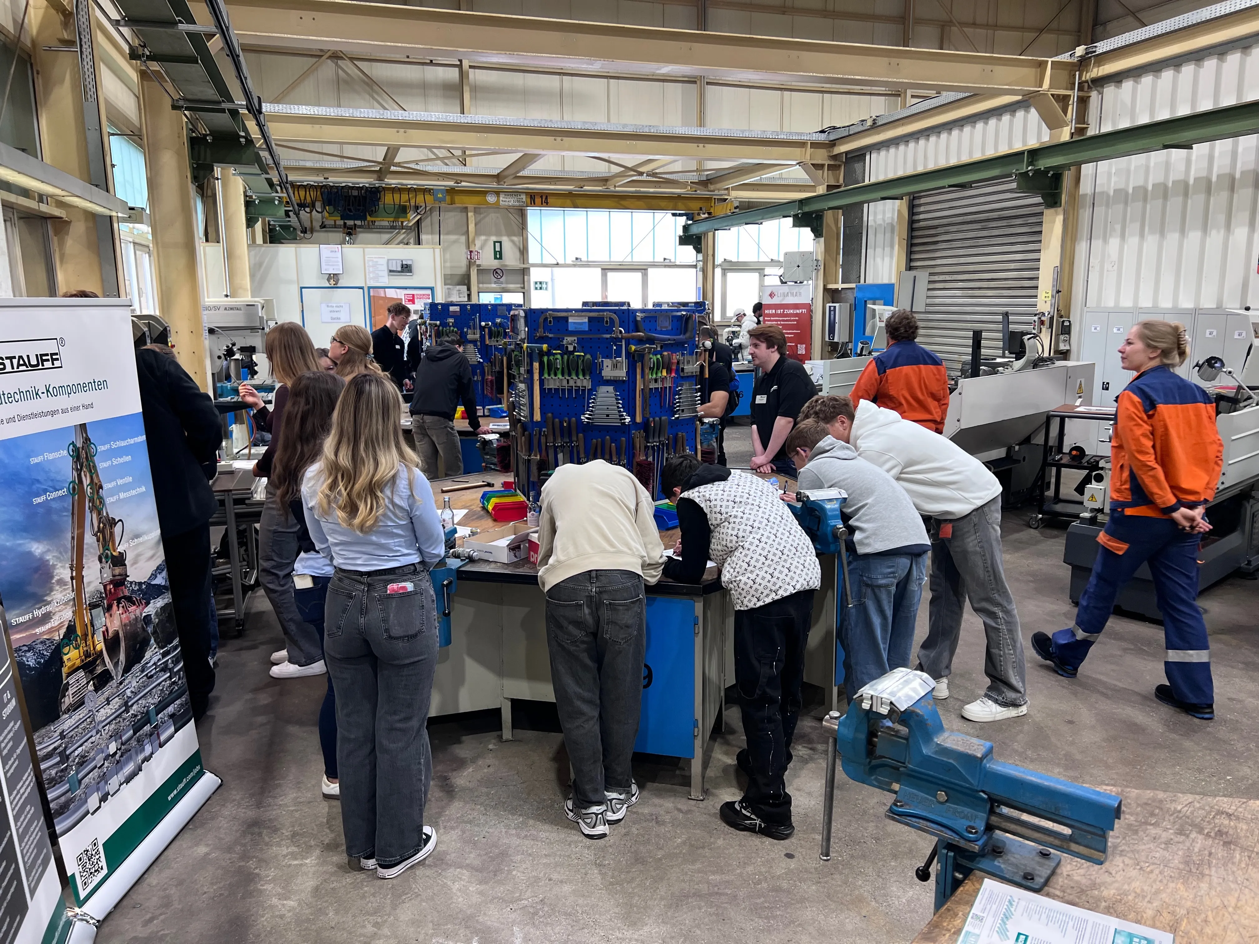 Eine Gruppe Jugendlicher steht in einer Werkstatt und beobachtet einen Mann, der an einem Metallstück arbeitet. Im Hintergrund sind Werkzeuge und Schränke zu sehen.