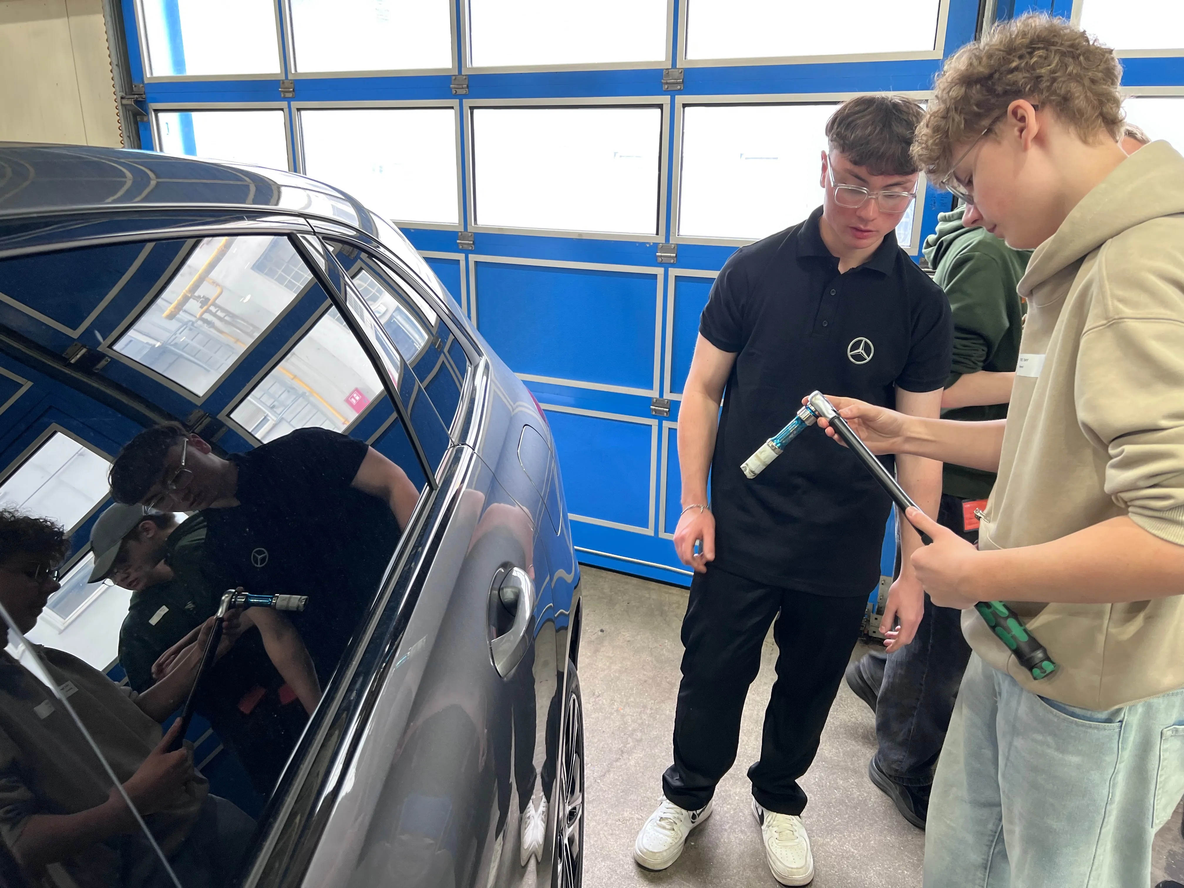 Zwei junge Männer untersuchen gemeinsam die Lackierung einer dunklen Limousine, während ein weiterer junger Mann im Hintergrund arbeitet. Sie nehmen an einem TecDay der SIHK teil und lernen gewerblich-technische Berufe kennen.