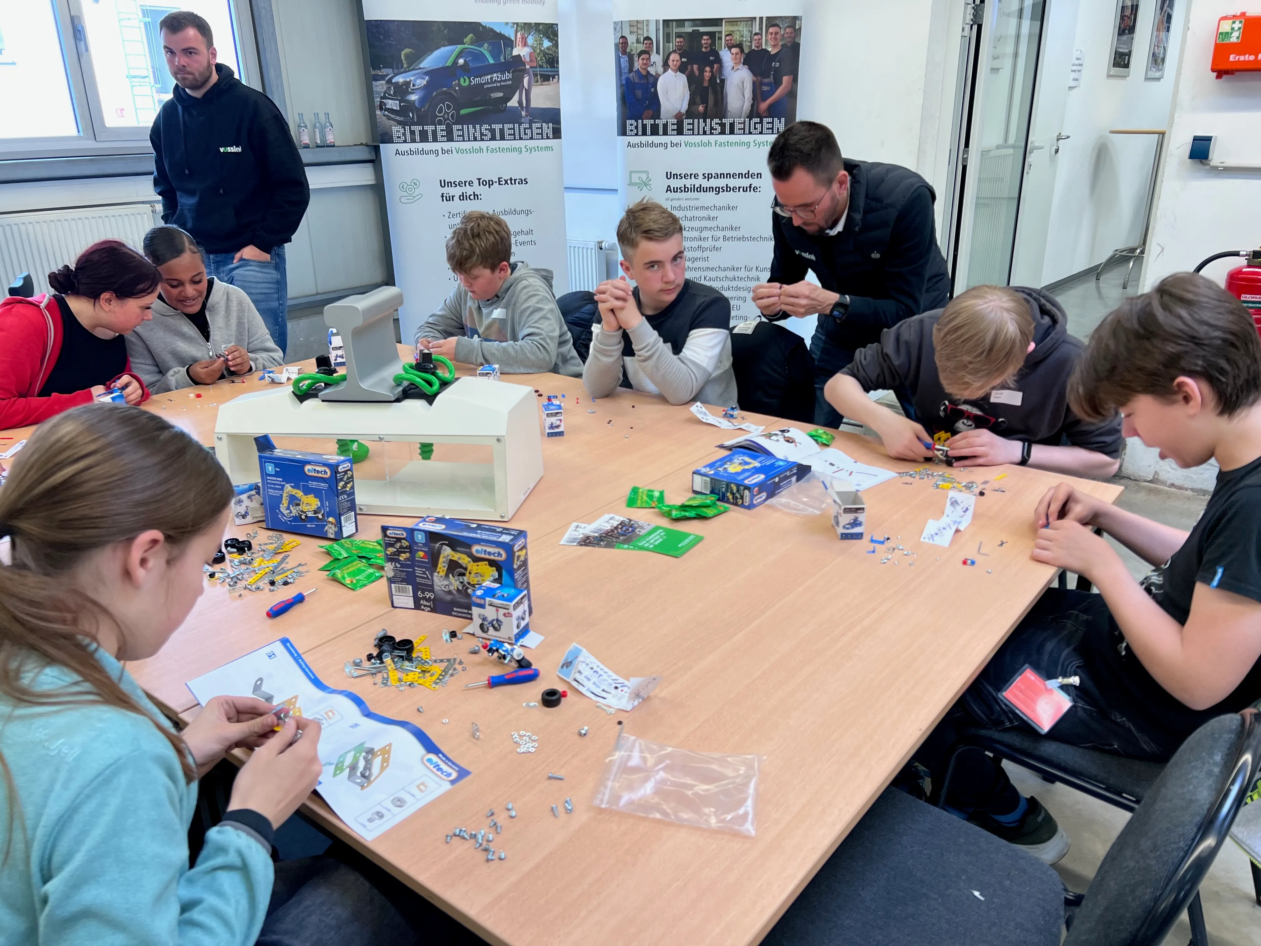 Schülergruppen sitzen an Tischen und arbeiten konzentriert mit kleinen Bauteilen, vermutlich im Rahmen eines Technik-Workshops. Im Hintergrund hängen Plakate mit Informationen zur Berufsausbildung.